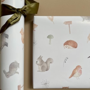 May include: Two rolls of white wrapping paper with a woodland animal theme. The paper features illustrations of squirrels, hedgehogs, rabbits, birds, mushrooms, and other woodland elements. The paper is tied with a green ribbon.