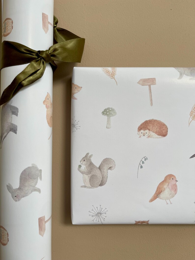 May include: Two rolls of white wrapping paper with a woodland animal theme. The paper features illustrations of squirrels, hedgehogs, rabbits, birds, mushrooms, and other woodland elements. The paper is tied with a green ribbon.