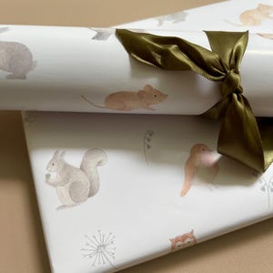 May include: White wrapping paper with a woodland animal theme. The paper features illustrations of a squirrel, a mouse, a bird, an owl, and a rabbit. The paper is rolled up and tied with a green ribbon.