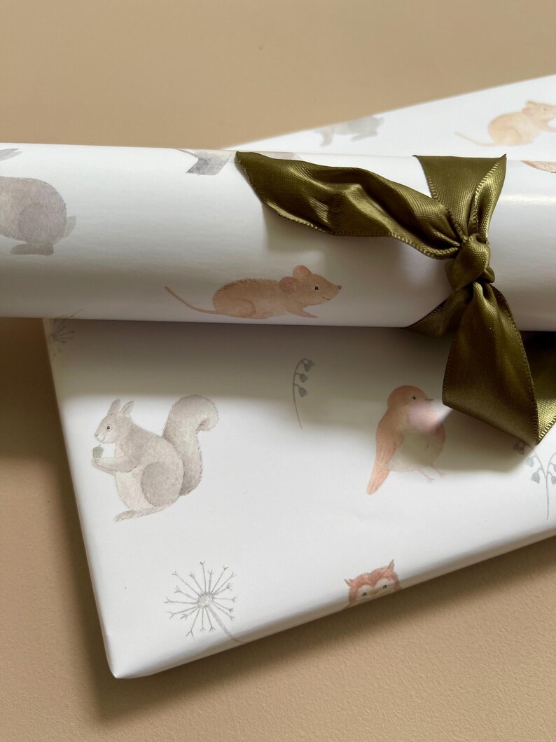 May include: White wrapping paper with a woodland animal theme. The paper features illustrations of a squirrel, a mouse, a bird, an owl, and a rabbit. The paper is rolled up and tied with a green ribbon.