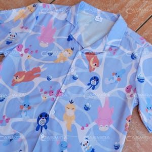 May include: A light blue button-up shirt with a cartoon print of various Pokemon characters. The shirt has a white background with a blue pattern.
