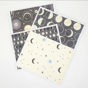 May include: Four cards with different moon and star patterns. The cards are black with white and gold accents. One card has a cream background with blue and gold stars and moons.
