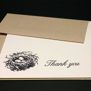 May include: A white thank you card with a black and white illustration of a bird's nest with eggs. The card is folded in half and has a brown envelope.