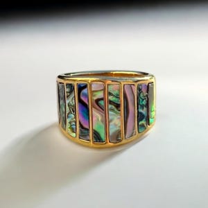 Puede incluir: Un anillo de oro con una banda ancha con incrustaciones de concha de abulón iridiscente. La concha está cortada en formas rectangulares y engastada en un marco de oro.