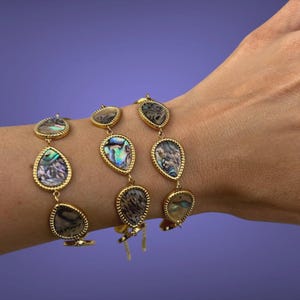 May include: Three gold-toned bracelets with teardrop-shaped abalone shell pendants. The iridescent shells are set in gold bezels with a textured edge. The bracelets are displayed on a purple background.
