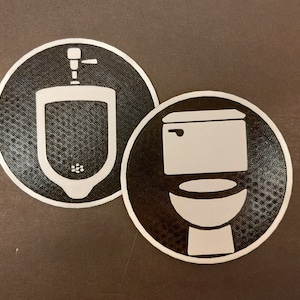 May include: Two circular restroom signs. One features a white outline of a urinal, the other a white outline of a toilet. Both are set against a black background with a dotted texture and a silver border.