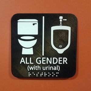 May include: Black and white sign with a toilet and urinal icon. The sign reads "ALL GENDER (with urinal)" and has braille below the text.