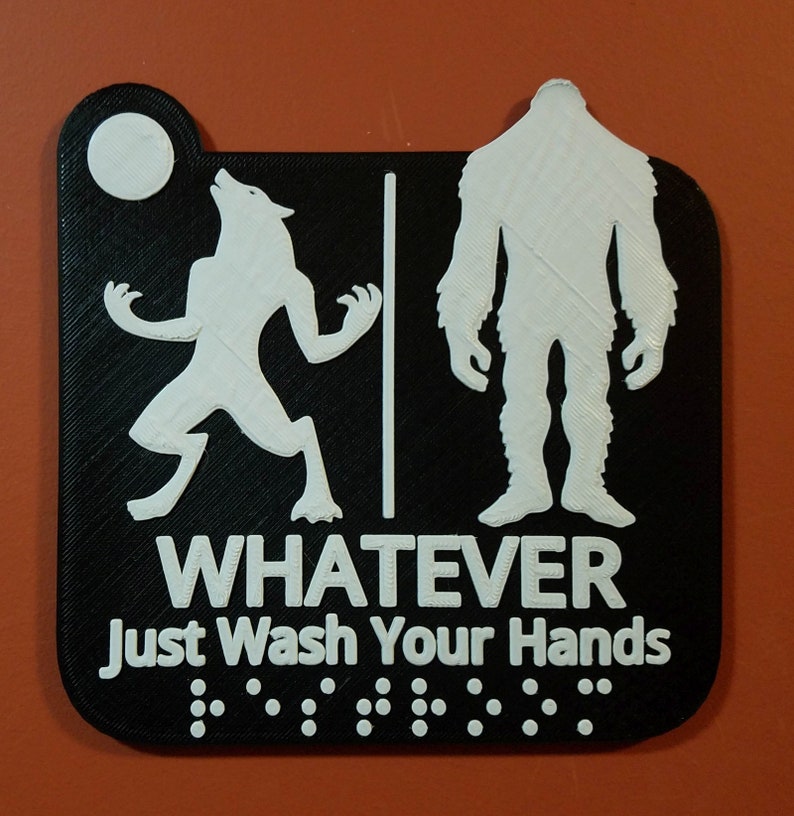 May include: Black and white bathroom sign with a werewolf and a bigfoot silhouette. The sign reads "Whatever Just Wash Your Hands" with braille dots below the text.