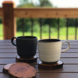 May include: Two ceramic mugs, one black and one white, sitting on wooden coasters. The mugs are on a wooden table with a blurred background of a wooden deck and green trees.