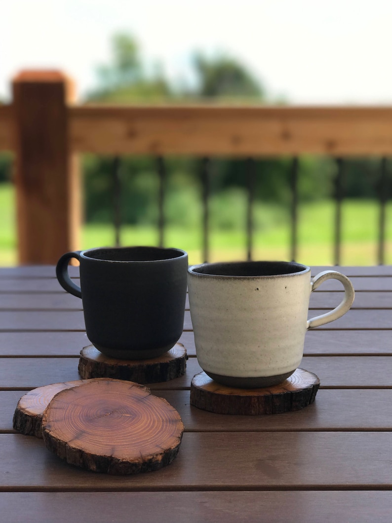May include: Two ceramic mugs, one black and one white, sitting on wooden coasters. The mugs are on a wooden table with a blurred background of a wooden deck and green trees.