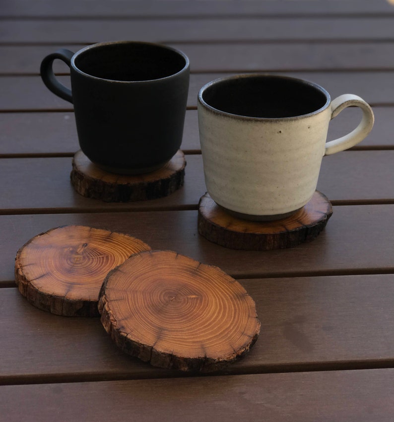 May include: Two ceramic mugs, one black and one white, are sitting on wooden coasters. The coasters are made from tree rounds and have a natural wood finish.