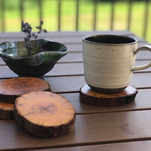 May include: Three wooden coasters with a natural wood grain finish, stacked on top of each other. A white ceramic mug with a dark brown rim sits on top of the coasters. A small, dark green ceramic bowl sits on the table next to the mug.