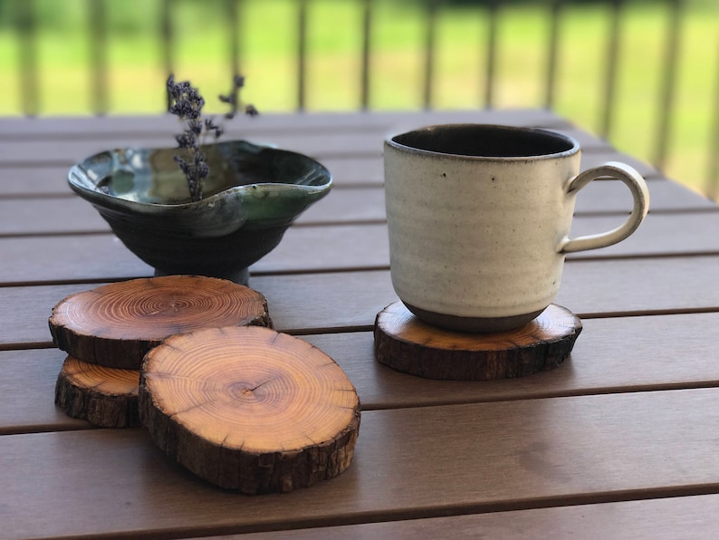 May include: Three wooden coasters with a natural wood grain finish, stacked on top of each other. A white ceramic mug with a dark brown rim sits on top of the coasters. A small, dark green ceramic bowl sits on the table next to the mug.