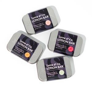 May include: Four silver tins of lotion bars, each with a different scent. The tins are labeled "Zerra & Co. Lotion Bar" and have a small circle with the scent name on each. The scents are Rose Petal, Orange Blossom, Lavender, and Cucumber.