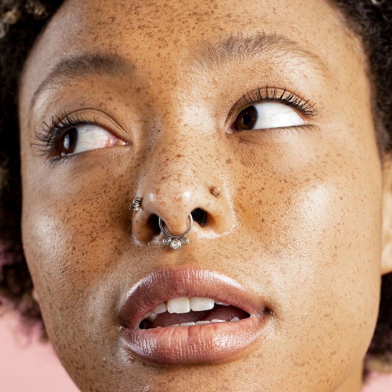 Puede incluir: Primer plano del rostro de una persona con m&uacute;ltiples piercings faciales. El piercing en el tabique es un anillo plateado con tres gemas transparentes. La piel tiene pecas y los labios son brillantes.