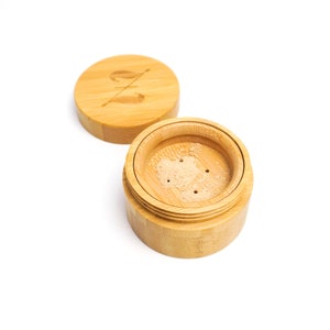 May include: A wooden container with a lid, containing a light brown powder. The container has a small logo on the lid.