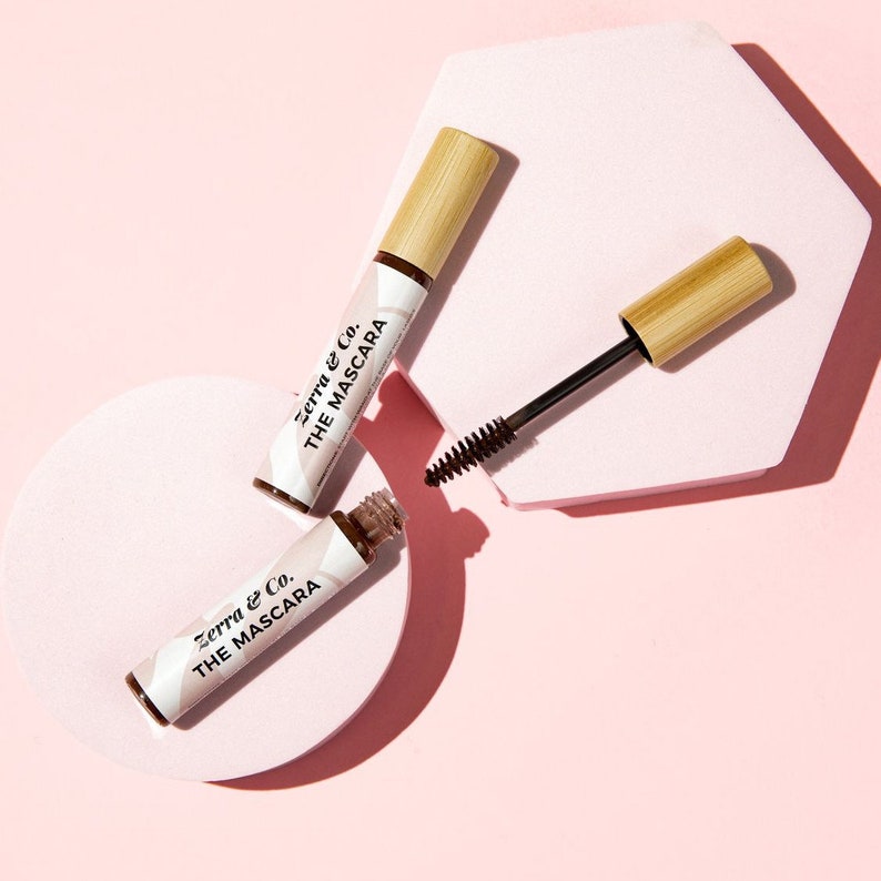 Puede incluir: Tres tubos de m&aacute;scara de pesta&ntilde;as con tapas de bamb&uacute; y el texto "Zerra & Co. THE MASCARA" impreso en las etiquetas blancas. Un tubo est&aacute; abierto, revelando el cepillo. El fondo es rosa claro con formas geom&eacute;tricas.