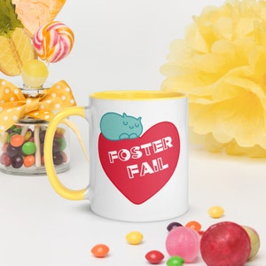 Peut inclure: Une tasse en céramique blanche avec un bord jaune. La tasse présente un cœur rouge avec le texte "Foster Fail" en blanc. Un chat bleu dort sur le cœur.