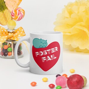 Peut inclure: Une tasse en céramique blanche avec un motif de cœur rouge. Le cœur porte le texte "Foster Fail" en lettres blanches. Un chat bleu dort sur le cœur.