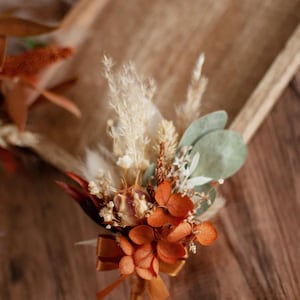 Pode incluir: Uma flor de lapela composta por pétalas de hortênsia laranja secas, erva-das-pampas bege, alpista branca e folhas de eucalipto verdes. O arranjo é atado com uma pequena fita de cetim laranja-escuro na base dos caules.