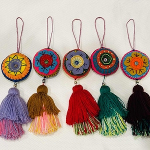 Op de afbeelding: Een verzameling van vijf kleurrijke, ronde stoffen ornamenten met geborduurde bloemmotieven. Elk ornament heeft een kwast in een andere kleur, waaronder paars, bruin, rood, groen en donkerbruin. De ornamenten zijn opgehangen aan een paars touwtje.