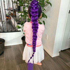 May include: A young person with long, braided purple hair, adorned with gold accents. The braid extends down the back, showcasing the vibrant color. The person is wearing a pink shirt, a white skirt with small dots, and purple leggings.