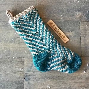 May include: A teal and white striped knitted stocking with a chevron pattern. The stocking has a brown knitted cuff and a loop at the top. The stocking is lying on a wooden surface with a wooden tag that says "@dr.knits".