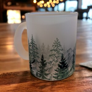 May include: A white frosted mug with a watercolor design of a forest scene. The mug has a handle and is sitting on a wooden table.