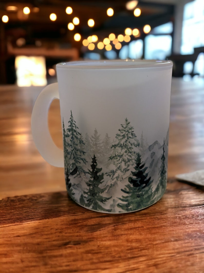 May include: A white frosted mug with a watercolor design of a forest scene. The mug has a handle and is sitting on a wooden table.