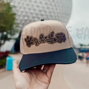 May include: Tan and navy blue baseball cap with a black brim. The cap features a brown design of Mickey Mouse gloves, Minnie Mouse bow, a wizard hat, and Mickey Mouse face. The background includes a large white sphere and the text "PAIRED EARS".