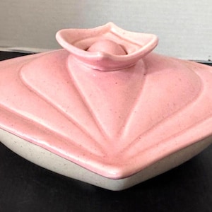 May include: A vintage pink ceramic dish with a lid. The lid has a stylized flower-like design with a small knob on top. The dish has a speckled, off-white base and a pink rim.