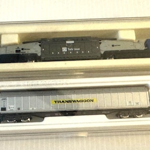 May include: Two model train cars in clear plastic display cases. The top car is a gray and blue crane unit with the text Transwaggon. The bottom car is a silver freight wagon with the yellow and black text Transwaggon printed on the side.