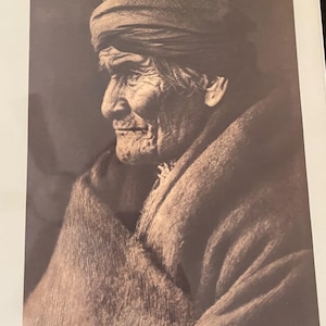 Op de afbeelding: Sepia-getint portret van een oudere persoon in profiel, met een hoofdband en gehuld in een deken. De afbeelding benadrukt het verweerde gezicht van de persoon, met zichtbare rimpels en een contemplatieve uitdrukking. De foto is een studie in textuur en licht.