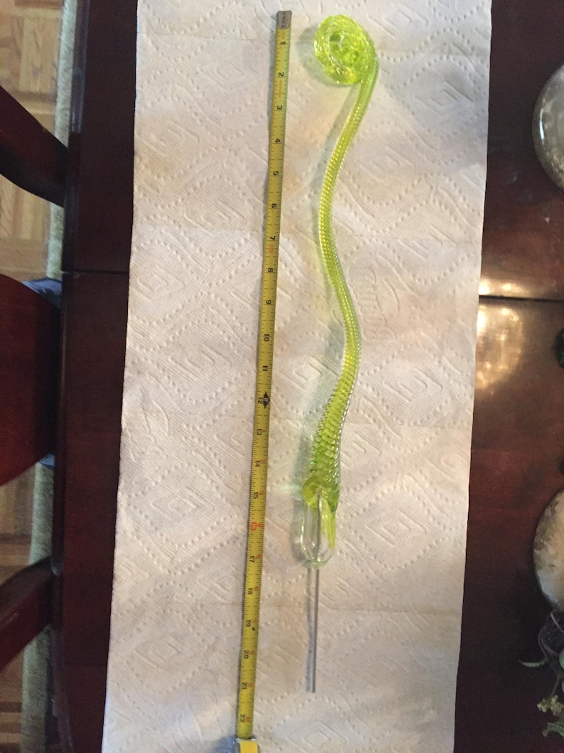 May include: A bright green glass floral stem with a curled top. The stem is about 18 inches long and is made of textured glass. The stem is on a white surface with a ruler next to it.