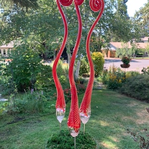May include: Three red glass sculptures with spiral tops are arranged in a pot with green moss. The sculptures are tall and slender, resembling stylized flowers.