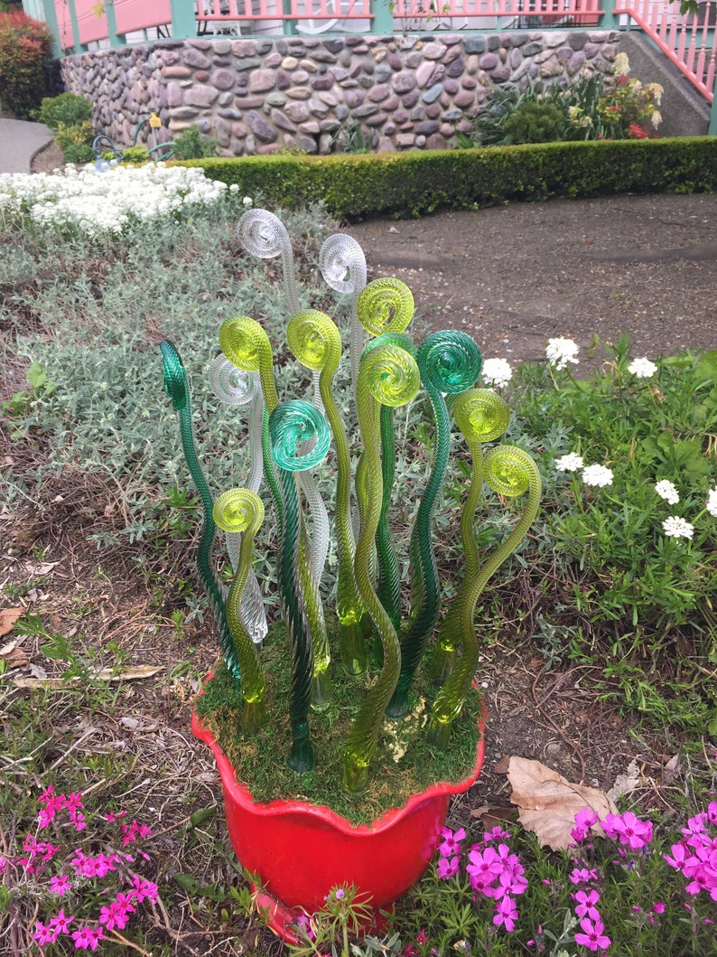 May include: A red ceramic planter with a green moss-like lining and a cluster of glass fern sculptures in shades of green and clear.