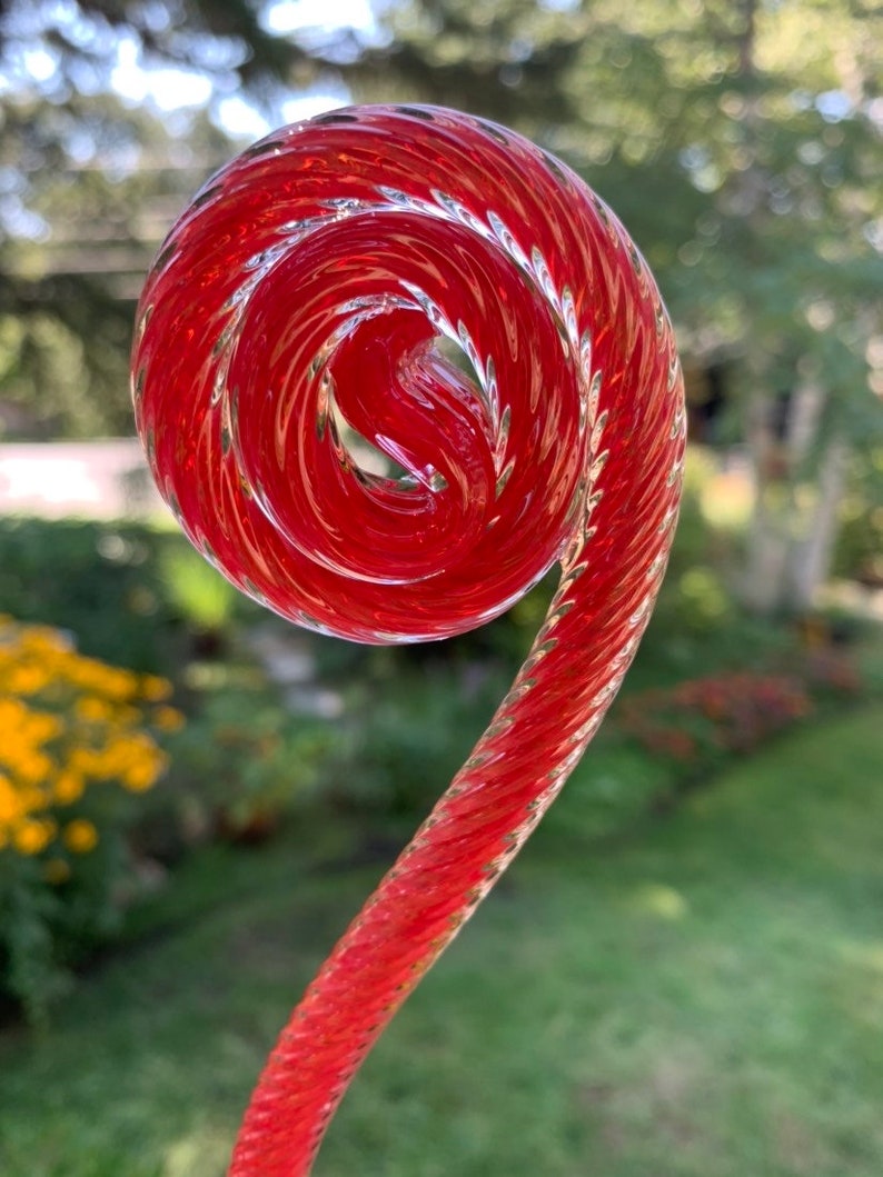 May include: A red glass spiral, twisted and curled, with a clear glass finish. The spiral is a decorative element for a garden or home.