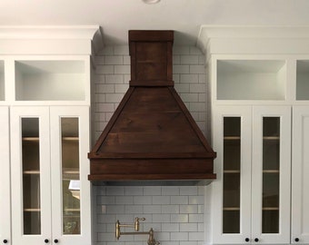 May include: A stainless steel range hood with a dark wood canopy, a gas hob, and a stainless steel oven. The range hood is mounted above the hob and is attached to the wall with white tiles.