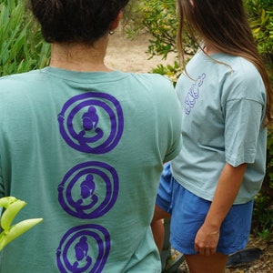 May include: Two people wearing light blue t-shirts with purple graphic designs on the back. The person on the left has a bun in their hair and is wearing blue jeans. The person on the right is wearing blue shorts.
