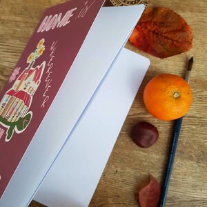 以下が含まれることがあります： 「Home is wherever」というテキストと、屋根の上に亀がいる家の絵が描かれたピンクのノート。ノートが開いていて、内側のページは白いです。