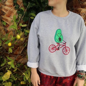 Può includere: Una felpa grigia con una grafica di un avocado che guida una bicicletta. L'avocado ha un viso sorridente e gli occhi chiusi.