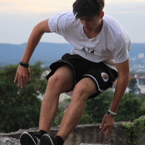 May include: A person wearing a white t-shirt and black shorts with a white stripe and a logo, performing a parkour move. The background features a cityscape and a cloudy sky.