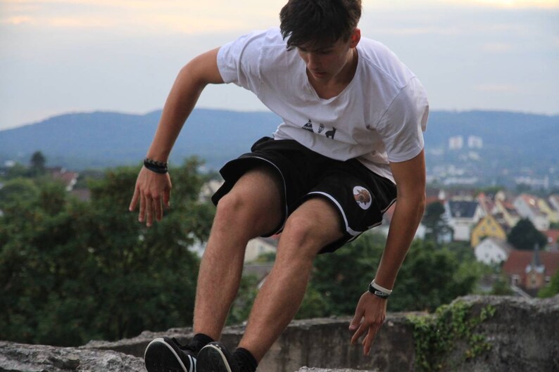 May include: A person wearing a white t-shirt and black shorts with a white stripe and a logo, performing a parkour move. The background features a cityscape and a cloudy sky.