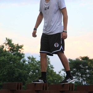 May include: A person wearing a white t-shirt and black shorts with a white trim and a logo. The person is wearing black socks and black sneakers with white stripes. The background includes green trees and a colorful sky.