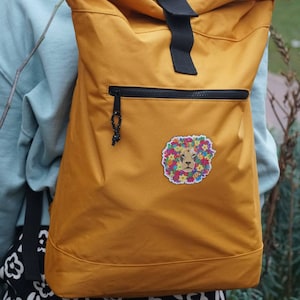 May include: A mustard yellow backpack with a black zipper and a floral lion patch. The backpack has a roll-top closure and adjustable straps.