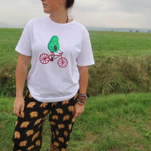 Pode incluir: Camiseta branca com um abacate verde andando de bicicleta. O abacate est&aacute; sorrindo e tem uma bicicleta vermelha com rodas pretas.