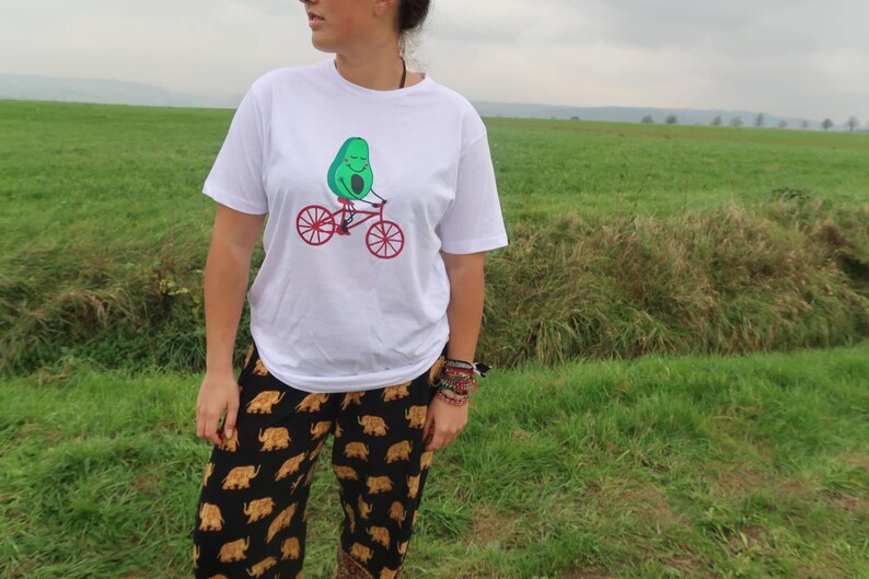 Pode incluir: Camiseta branca com um abacate verde andando de bicicleta. O abacate est&aacute; sorrindo e tem uma bicicleta vermelha com rodas pretas.