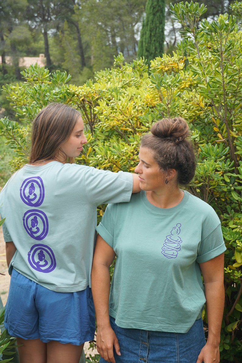 May include: Two people wearing teal blue t-shirts with purple designs. The design on the left shirt is three stacked circles with a person in each circle. The design on the right shirt is a single circle with a person inside and stacked circles around it.