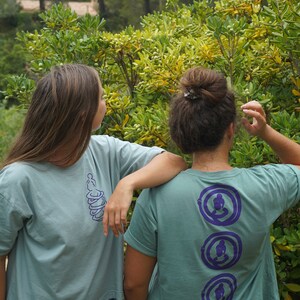 May include: Two people wearing teal blue t-shirts with purple designs. The person in the back has three circular designs with a stylized figure in each. The person in the front has a single design of a stylized figure.