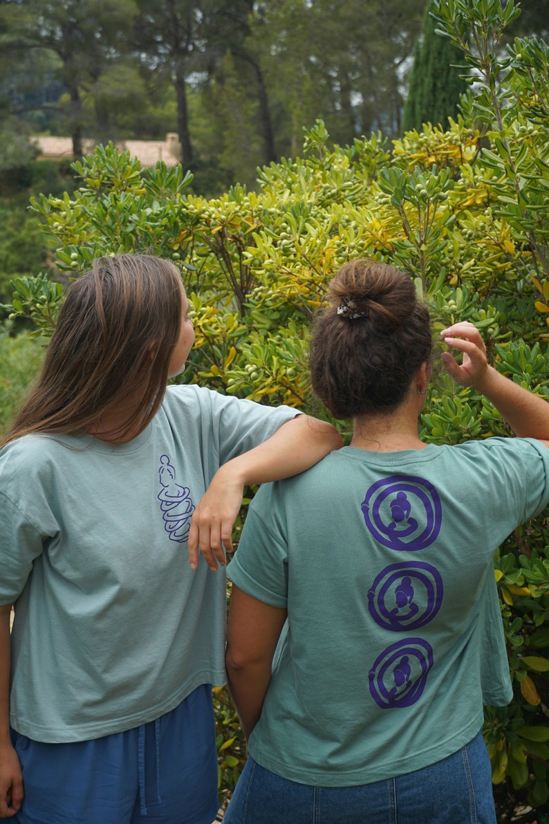 May include: Two people wearing teal blue t-shirts with purple designs. The person in the back has three circular designs with a stylized figure in each. The person in the front has a single design of a stylized figure.